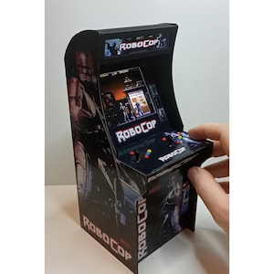 May include: Miniature black arcade cabinet with Robocop branding. The cabinet has a screen showing a game scene, with buttons and a joystick. The sides and top also display the Robocop logo and imagery.