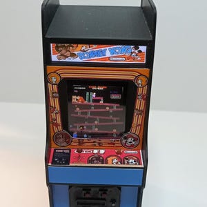 Può includere: Un cabinato arcade in miniatura di Donkey Kong con cornice nera, pannello inferiore blu e uno schermo che mostra il gioco classico. La console include un joystick, pulsanti di controllo e grafiche colorate con i loghi Donkey Kong e Nintendo.