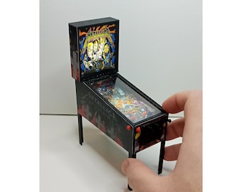 May include: Miniature pinball machine with a black and red design. The backboard features the band Metallica's logo and artwork. The machine has a glass-covered playfield and is supported by four black legs. A hand is visible, holding the machine.