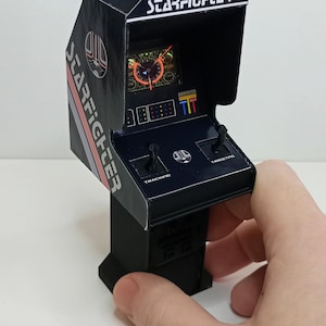 Może przedstawiać: Miniaturowa czarna gra arkadowa ze słowem "STARFIGHTER" na górze. Gra ma ekran wyświetlający kolorową grafikę, dwa joysticki oraz słowa "TRACKING" i "TARGETING". Gra arkadowa jest trzymana w dłoni.
