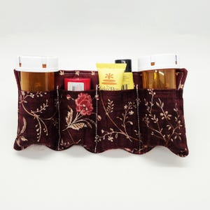 May include: A burgundy fabric organizer with four pockets, featuring a floral pattern in cream and gold. The organizer holds two amber prescription bottles, a red container, and two yellow and black tubes. The edges have a scalloped design.