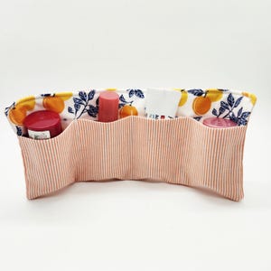 May include: A fabric organizer with a peach and white striped exterior and a citrus fruit and floral patterned interior. The organizer has multiple pockets holding various cosmetic items, including lipsticks and creams.
