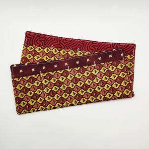 May include: Two fabric pen cases. The cases are burgundy, yellow, and red with a floral pattern. The top case has a burgundy swirl pattern. The bottom case has a burgundy band with white leaf shapes. White stitching is visible.