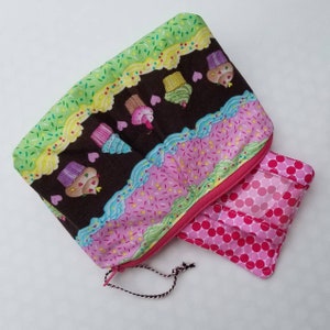 May include: A pink and green zippered pouch with a brown fabric lining and a cupcake pattern. The pouch has a clear plastic window on the front and a pink polka dot fabric lining.