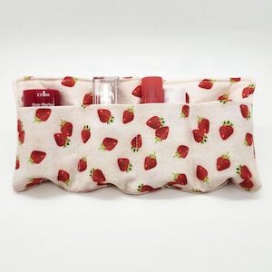 May include: A fabric organizer with a strawberry pattern, holding various cosmetic items. The organizer is beige with a repeating pattern of red strawberries. The pockets hold lipsticks and other beauty products.