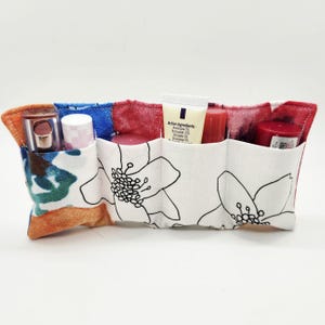May include: A fabric cosmetic organizer with multiple pockets, holding various beauty products. The organizer features a floral print on a white background, with colorful accents of blue, red, and orange. The organizer is filled with lipsticks, lip balms, and a tube of cream.