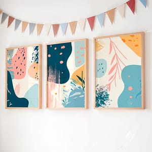 Pode incluir: Três impressões de arte abstrata emolduradas com formas coloridas em rosa, azul e amarelo. As impressões estão em molduras de madeira clara e penduradas em uma parede branca. Uma guirlanda de bandeiras triangulares está pendurada acima das impressões.