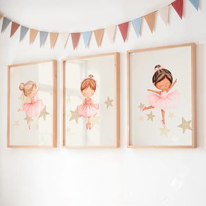 以下が含まれることがあります： ピンクのチュチュと金色の星が描かれたバレリーナの水彩画イラストが特徴の、額装された3つのプリント。プリントは明るい木製のフレームに入っており、白い壁に掛けられています。カラフルなバナーがプリントの上に吊り下げられています。