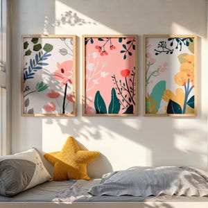 Pode incluir: Três impressões de arte floral emolduradas. Cada impressão apresenta um arranjo floral diferente com flores rosa, verdes e amarelas em um fundo branco ou rosa. As molduras são de cor de madeira clara. Uma almofada amarela em forma de estrela e outras almofadas estão em primeiro plano.