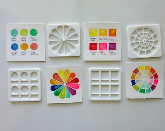 May include: Eight white plastic watercolor palettes with different layouts and sizes. The palettes are arranged in two rows of four. Some of the palettes have watercolor paint swatches in them, showing a variety of colors.