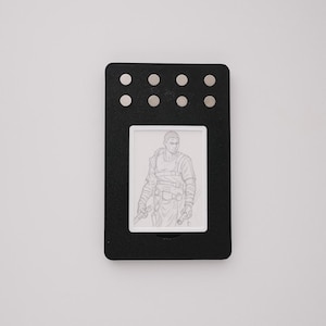 May include: A rectangular black display frame featuring eight small silver circular accents arranged in two rows at the top. Centered within the frame is a white rectangular insert containing a detailed black and white line drawing of a person.
