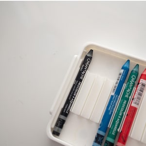 Peut inclure: Un &eacute;tui en plastique blanc contenant quatre crayons de couleur. Les crayons sont noir, bleu, vert et rouge. Les crayons sont &eacute;tiquet&eacute;s "Caran d'Ache Neocolor II Crayons aquarellables".