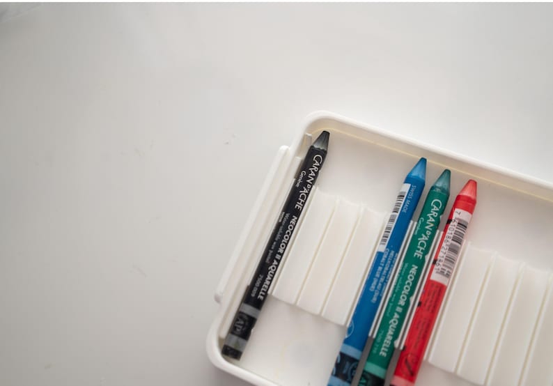Peut inclure: Un &eacute;tui en plastique blanc contenant quatre crayons de couleur. Les crayons sont noir, bleu, vert et rouge. Les crayons sont &eacute;tiquet&eacute;s "Caran d'Ache Neocolor II Crayons aquarellables".