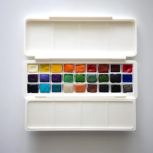 May include: A white plastic watercolor paint set with 24 individual pans of paint. The pans are arranged in a grid pattern and are filled with a variety of colors, including red, blue, green, yellow, and orange.