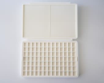 Könnte beinhalten: Eine weiße Plastik-Farbpalette mit 48 leeren Vertiefungen. Die Palette ist geöffnet und der Deckel liegt flach auf der Oberfläche.