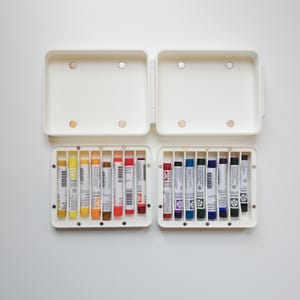May include: A white plastic storage case with two open compartments containing fourteen cylindrical oil sticks. The sticks are arranged in individual slots and feature labels with various colors including yellow, orange, red, blue, purple, and black.