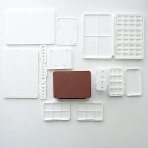 May include: A collection of white plastic art supplies, including palettes, trays, and a brown rectangular box, arranged on a white surface. The image suggests a studio or workspace with various art materials.