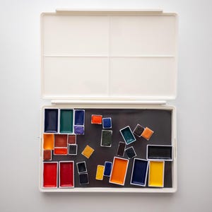 Pode incluir: Um conjunto de tinta de aquarela de plástico branco com uma bandeja preta contendo uma variedade de cores, incluindo azul, verde, vermelho, laranja e amarelo.