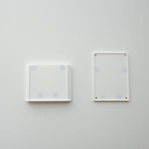 May include: Two white rectangular objects with rounded corners are displayed against a white background. One object is a tray with a raised edge, and the other is a flat, rectangular frame. Both have small, dark circular details.