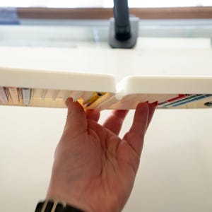 Peut inclure: Une bo&icirc;te de rangement en plastique blanc avec plusieurs compartiments, remplie de petits objets color&eacute;s. La bo&icirc;te est tenue ouverte par une main.