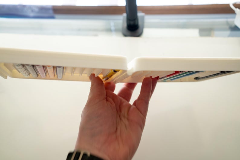 Peut inclure: Une bo&icirc;te de rangement en plastique blanc avec plusieurs compartiments, remplie de petits objets color&eacute;s. La bo&icirc;te est tenue ouverte par une main.