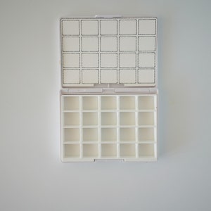 May include: White plastic watercolor paint palette with 24 empty wells. The palette is open and the lid is laying flat on top.