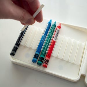 Peut inclure: Un &eacute;tui en plastique blanc avec un couvercle &agrave; charni&egrave;re, contenant un jeu de crayons de couleur. Les crayons sont dispos&eacute;s en rang&eacute;e, les pointes vers le haut. Les crayons sont bleu, vert, rouge et jaune. L'&eacute;tui porte une &eacute;tiquette indiquant "CARAN D'ACHE NEOCOLOR II AQUARELLE".