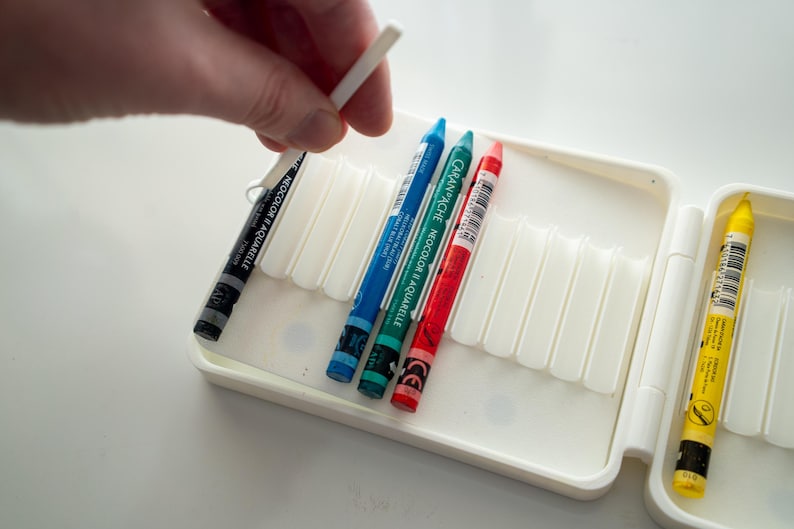 Peut inclure: Un &eacute;tui en plastique blanc avec un couvercle &agrave; charni&egrave;re, contenant un jeu de crayons de couleur. Les crayons sont dispos&eacute;s en rang&eacute;e, les pointes vers le haut. Les crayons sont bleu, vert, rouge et jaune. L'&eacute;tui porte une &eacute;tiquette indiquant "CARAN D'ACHE NEOCOLOR II AQUARELLE".