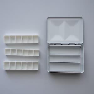 Könnte beinhalten: Eine weiße Aquarellfarbpalette mit offenem Deckel, drei Reihen einzelner Farbnäpfe und drei separaten Farbmischbereichen. Die Palette besteht aus Metall und Kunststoff.