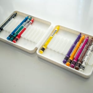 Peut inclure: Un &eacute;tui en plastique blanc avec deux rang&eacute;es de crayons de couleur. La rang&eacute;e du haut contient des crayons noirs, bleus, verts et rouges. La rang&eacute;e du bas contient des crayons jaunes, violets, oranges, roses, bruns et blancs.