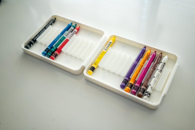 Peut inclure: Un &eacute;tui en plastique blanc avec deux rang&eacute;es de crayons de couleur. La rang&eacute;e du haut contient des crayons noirs, bleus, verts et rouges. La rang&eacute;e du bas contient des crayons jaunes, violets, oranges, roses, bruns et blancs.