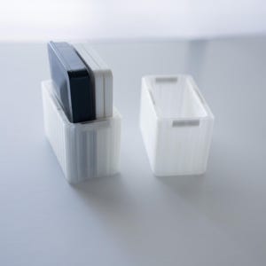 May include: Two white rectangular storage containers with a ribbed design. One container holds a black and a white rectangular object. The other container is empty. The containers are on a white surface.