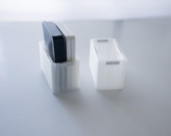 May include: Two white rectangular storage containers with a ribbed design. One container holds a black and a white rectangular object. The other container is empty. The containers are on a white surface.