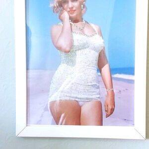 May include: A framed photograph of a person with blonde hair wearing a white, textured, sleeveless swimsuit and a beaded necklace. The person is standing on a sandy beach with a clear blue sky in the background, captured in a bright, sunny setting.