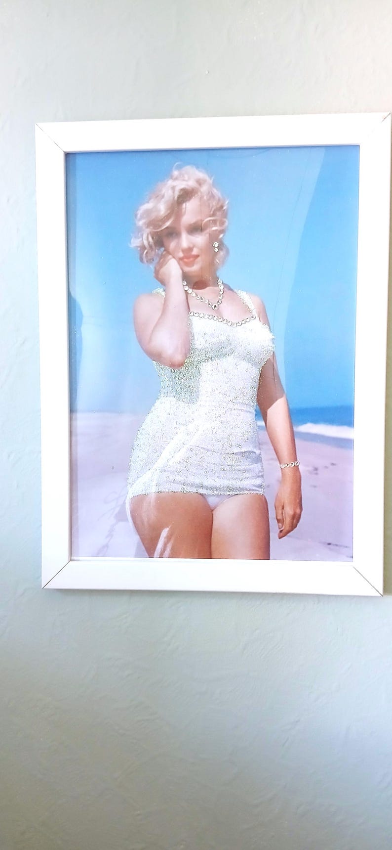 May include: A framed photograph of a person with blonde hair wearing a white, textured, sleeveless swimsuit and a beaded necklace. The person is standing on a sandy beach with a clear blue sky in the background, captured in a bright, sunny setting.