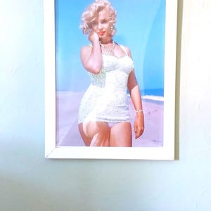 May include: A framed photograph featuring a blonde person with wavy hair wearing a white, textured, sleeveless swimsuit and a necklace. The person is standing on a sandy beach against a bright blue sky. The frame is white and rectangular.
