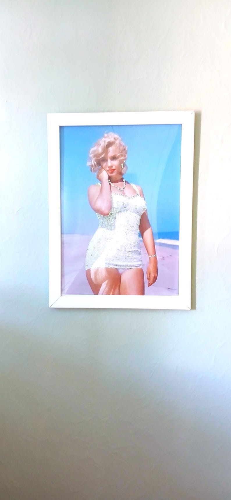 May include: A framed photograph featuring a blonde person with wavy hair wearing a white, textured, sleeveless swimsuit and a necklace. The person is standing on a sandy beach against a bright blue sky. The frame is white and rectangular.