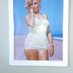 May include: A framed wall art piece featuring a glittery, sparkling image of a blonde person with wavy hair wearing a white one-piece swimsuit and a necklace, standing on a sandy beach against a bright blue sky, enclosed in a white frame.