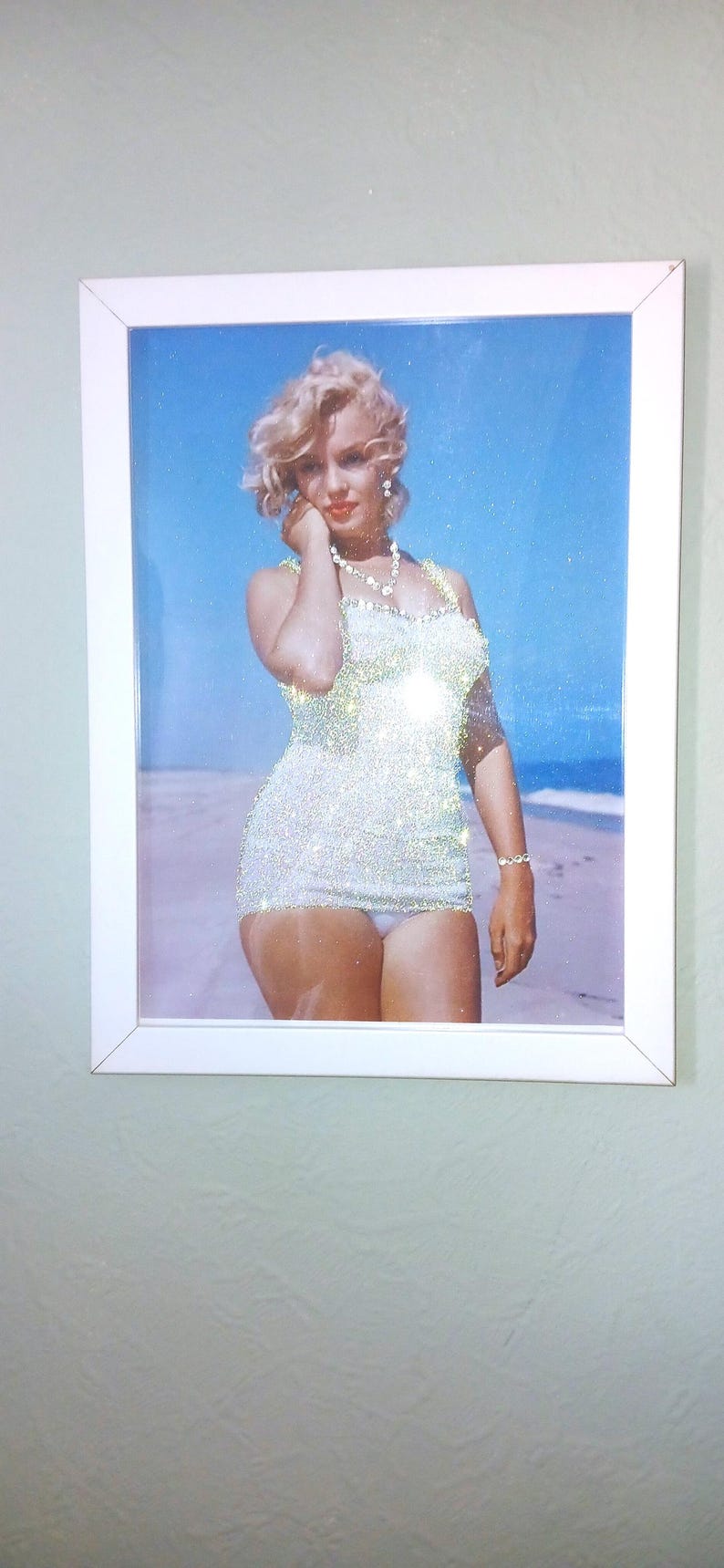 May include: A framed wall art piece featuring a glittery, sparkling image of a blonde person with wavy hair wearing a white one-piece swimsuit and a necklace, standing on a sandy beach against a bright blue sky, enclosed in a white frame.