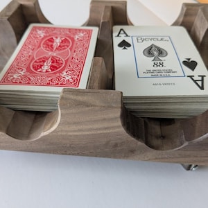 May include: A wooden playing card holder with two compartments, each containing a deck of cards. One deck shows a red patterned back, while the other shows the face of an Ace of Spades with the text Bicycle 88 The United States Playing Card Company.