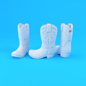 Op de afbeelding: Drie matwitte cowboylaarzen gerangschikt in een groep. Elke laars heeft een klassieke hoge schacht met decoratieve uitgesneden patronen, een puntige neus en een lage blokhak. De laarzen zijn uitgevoerd in een effen, uniforme witte kleur op blauw.