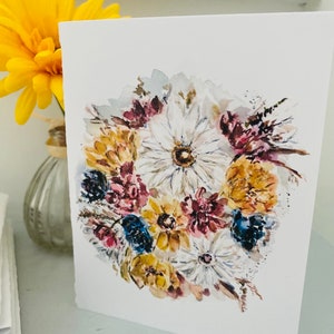 Puede incluir: Tarjeta de felicitaci&oacute;n blanca con una pintura de acuarela de un ramo floral. El ramo incluye flores blancas, amarillas, azules y burdeos. Una flor amarilla en un jarr&oacute;n de cristal es visible en el fondo.