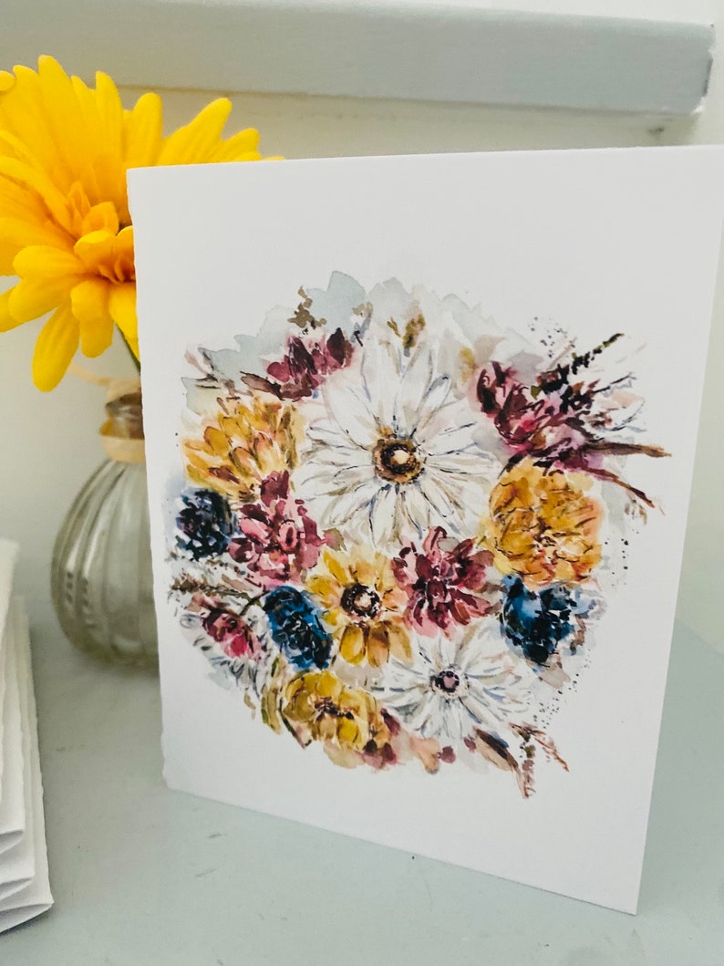 Puede incluir: Tarjeta de felicitaci&oacute;n blanca con una pintura de acuarela de un ramo floral. El ramo incluye flores blancas, amarillas, azules y burdeos. Una flor amarilla en un jarr&oacute;n de cristal es visible en el fondo.