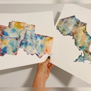K&ouml;nnte beinhalten: Zwei Aquarellbilder von Karten. Die erste Karte zeigt eine Stadt mit mehreren Stadtteilen, die in Blaut&ouml;nen, Rosa, Gelb und Braun gemalt sind. Die zweite Karte zeigt einen Bundesstaat oder eine Region, die in Blaut&ouml;nen, Gr&uuml;n und Braun gemalt ist. Beide Karten sind auf wei&szlig;em Papier.