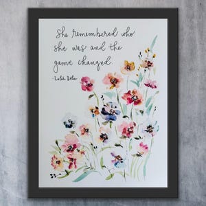 May include: Framed art print with a watercolour floral design in pink, red, blue, and yellow. The artwork includes the quote, "She remembered who she was and the game changed." by Lalah Delia.