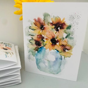 Puede incluir: Pintura a la acuarela de girasoles en un jarr&oacute;n azul sobre una tarjeta blanca. Las flores son amarillas, marrones y verdes. Una flor amarilla est&aacute; detr&aacute;s de la tarjeta. A la izquierda hay una pila de tarjetas plegadas.