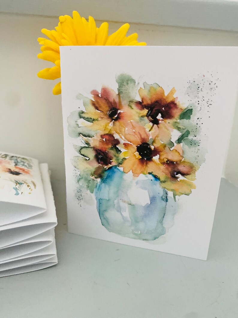 Puede incluir: Pintura a la acuarela de girasoles en un jarr&oacute;n azul sobre una tarjeta blanca. Las flores son amarillas, marrones y verdes. Una flor amarilla est&aacute; detr&aacute;s de la tarjeta. A la izquierda hay una pila de tarjetas plegadas.