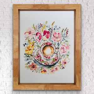 Puede incluir: Pintura a la acuarela de una taza y un platillo llenos de té, rodeados de un arreglo floral. La taza y el platillo tienen un estampado floral. La obra de arte está enmarcada en un marco de madera marrón claro.