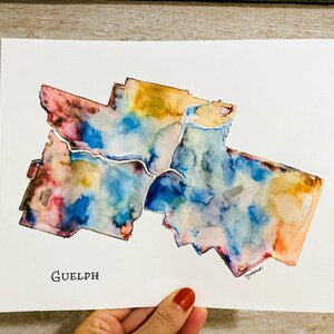 K&ouml;nnte beinhalten: Aquarellmalerei der Stadt Guelph, Ontario, Kanada. Die Karte ist in Blaut&ouml;nen, Rosa, Gelb und Orange gemalt. Der Stadtname "Guelph" ist unten auf der Karte in Schwarz geschrieben.