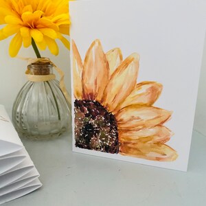 Puede incluir: Una tarjeta de felicitaci&oacute;n blanca con una pintura de acuarela de un girasol. El girasol tiene p&eacute;talos amarillos y un centro marr&oacute;n oscuro. Una flor amarilla en un jarr&oacute;n de cristal est&aacute; en el fondo.